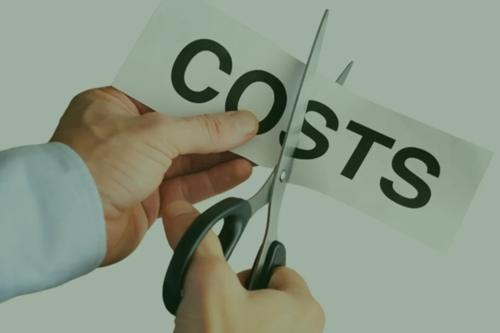 Male hand holding a note written "costs". With the other hand, the man cuts this note with scissors.